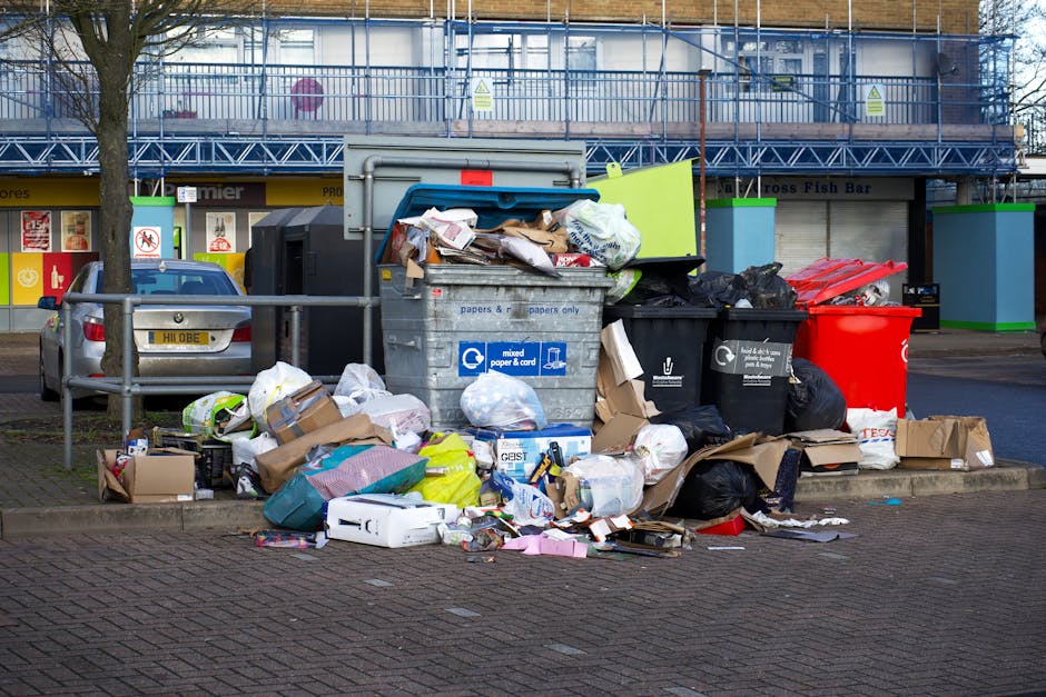An outdoor scene showing a cluttered collection of overflowing rubbish bins and scattered waste on a paved area in front of a retail building. The waste includes various types of discarded packaging, 