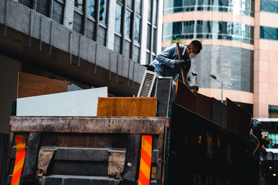 A man wearing a face mask, grey shirt, and dark trousers is standing inside the bed of a large, open-top black waste collection truck, which is parked outdoors in an urban environment. The truck conta