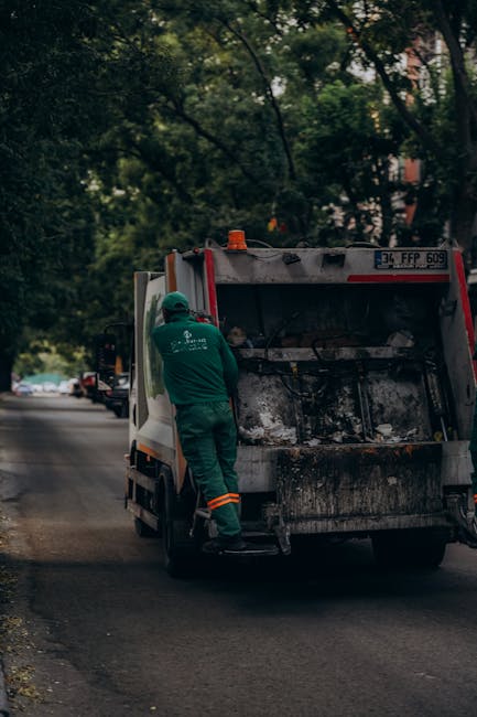 A man wearing a face mask, grey shirt, and dark trousers is standing inside the bed of a large, open-top black waste collection truck, which is parked outdoors in an urban environment. The truck conta