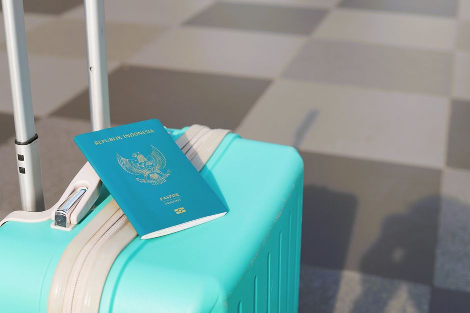 Close-up view of a light blue hard-shell suitcase with a cream-colored zipper and extendable handle, standing on a checkered pavement surface. Resting on top of the suitcase is a dark blue Indonesian 