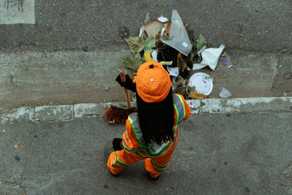 An individual dressed in high-visibility orange safety clothing and a matching orange cap is seen from an overhead perspective, sweeping a pavement with a long-handled broom made of wooden and bristle